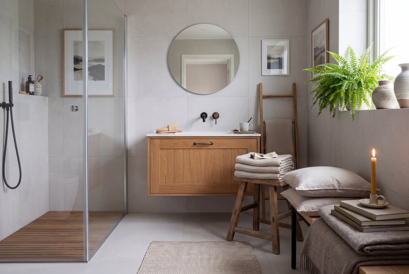 A Scandinavian bathroom with large-format pale tile, a wooden vanity, a round mirror and waffle-weave towels on a wooden ladder