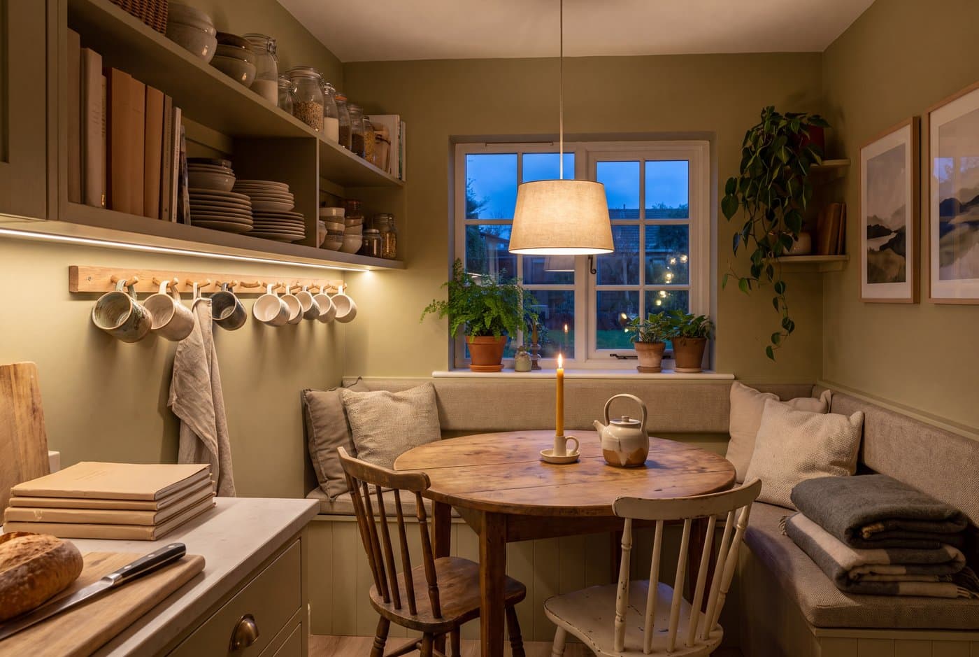A hygge kitchen with a small table and bench, warm painted cabinets, a wooden countertop and mugs on a hook rail